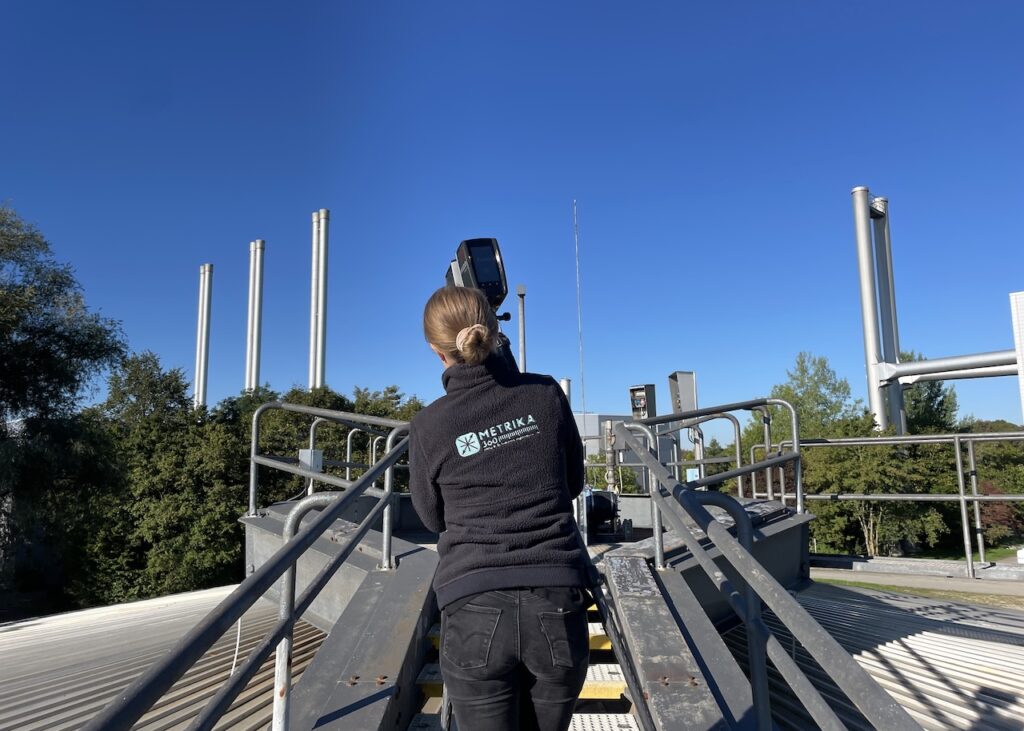 Surveyor with a scanner on a rooftop walkway, industrial stacks behind